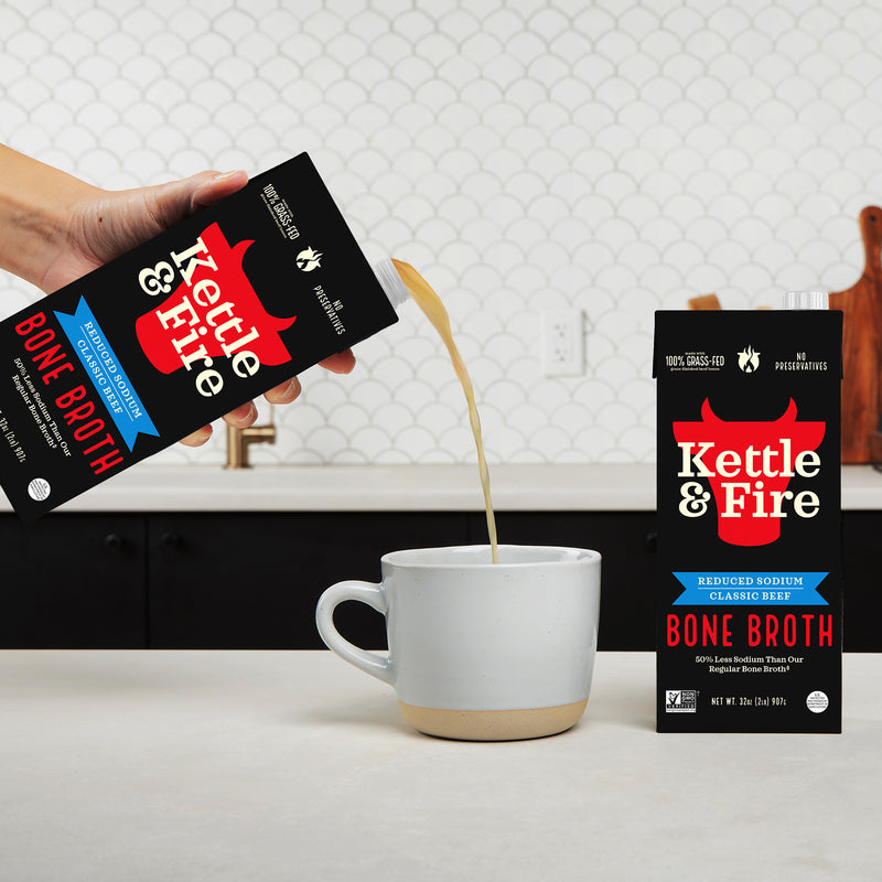Hand pouring Kettle & Fire reduced sodium beef bone broth from a black carton into a white ceramic mug.