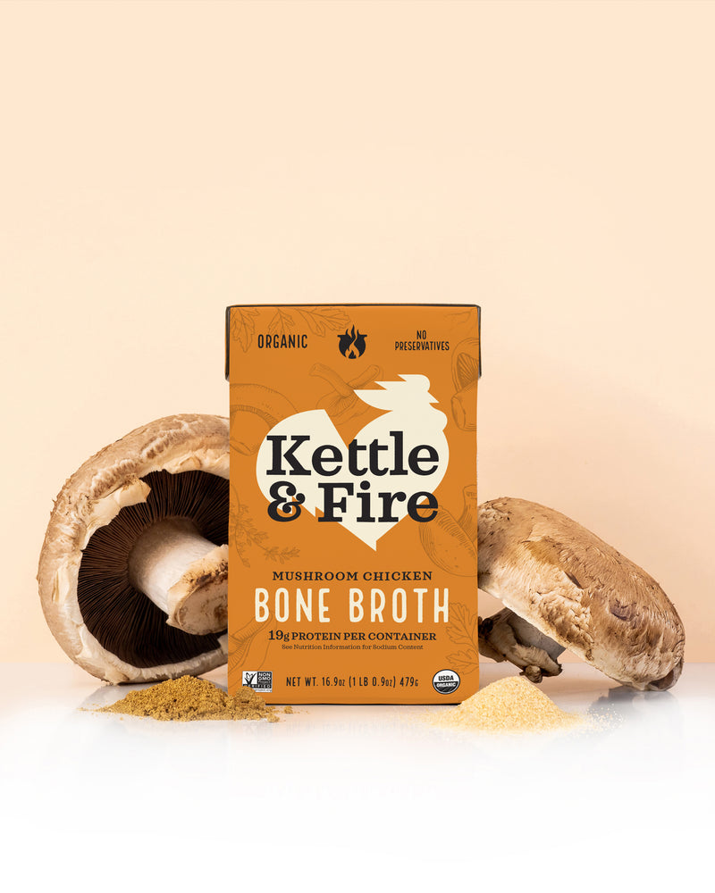 Kettle & Fire Mushroom Chicken Bone Broth carton surrounded by whole and sliced mushrooms on beige background.