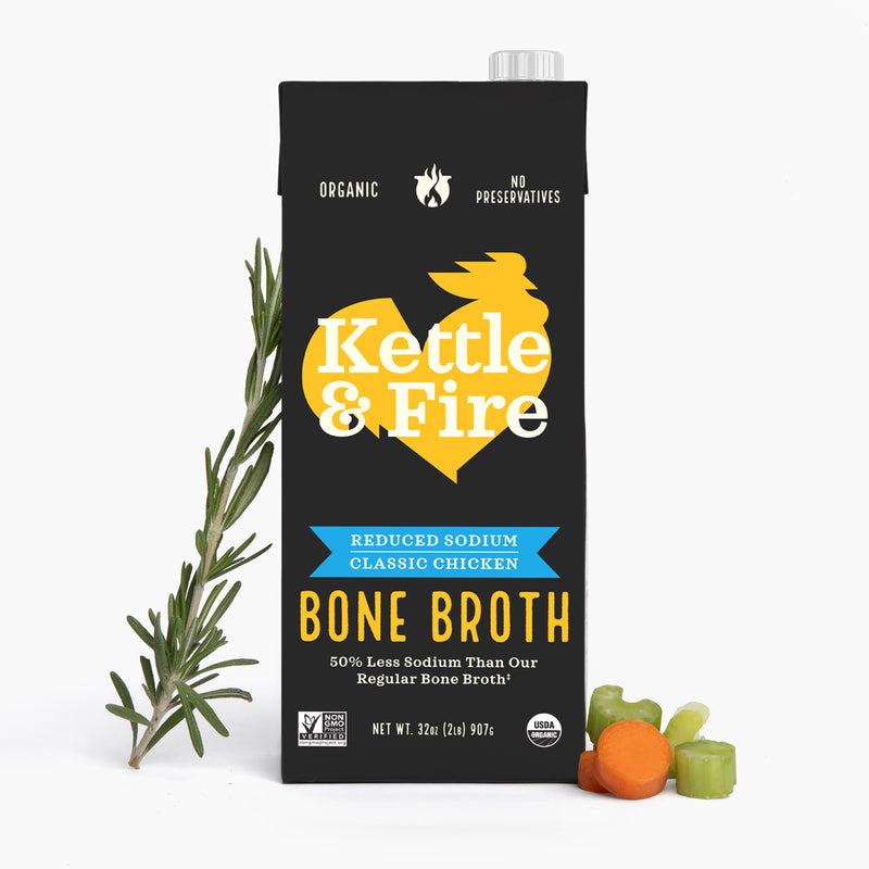 Black carton of Kettle & Fire reduced sodium chicken bone broth with rosemary sprigs and vegetables displayed beside it.