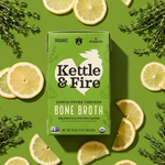 Kettle & Fire lemon thyme chicken bone broth carton surrounded by lemon slices and thyme sprigs on green background.